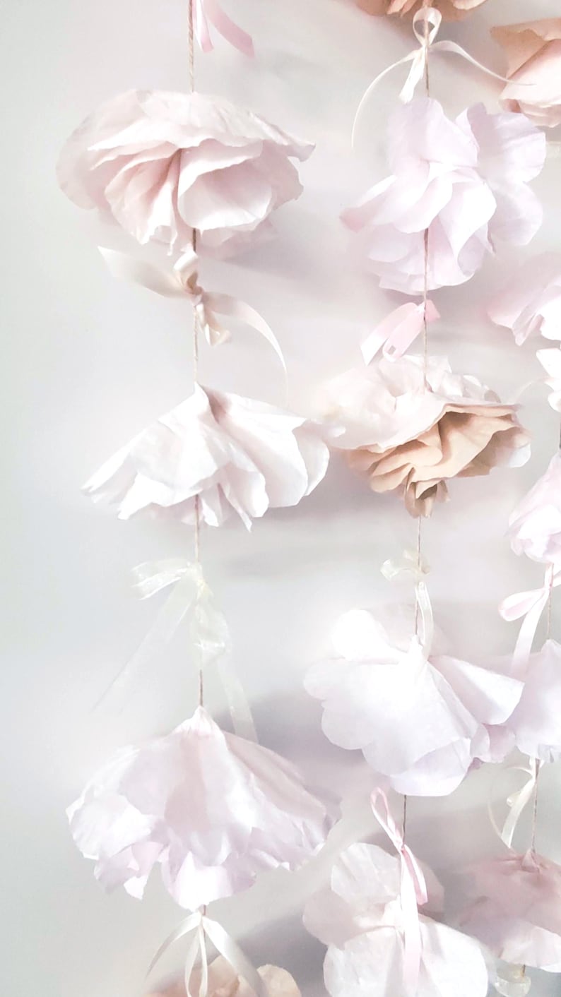 May include: A delicate garland of paper flowers in varying shades of pink and cream. The flowers are suspended by thin strings and adorned with small ribbons. The soft, layered petals create a romantic and airy aesthetic, perfect for event decor.