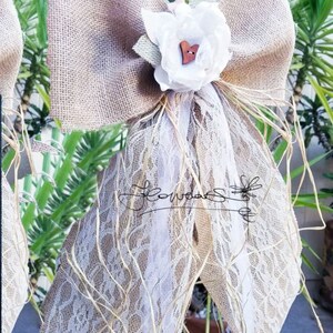 May include: A burlap bow with a white lace overlay and a white fabric flower with a wooden heart button in the center. The bow is tied with a brown ribbon and has a rustic, country feel.