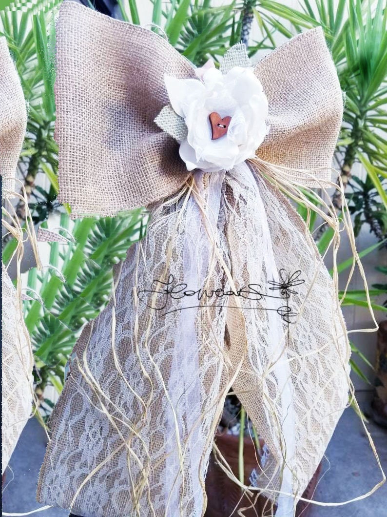 May include: A burlap bow with a white lace overlay and a white fabric flower with a wooden heart button in the center. The bow is tied with a brown ribbon and has a rustic, country feel.
