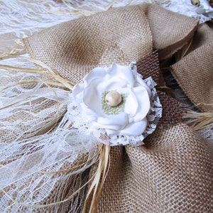 May include: A burlap and lace wedding ring pillow with a white fabric flower and a brown burlap bow.