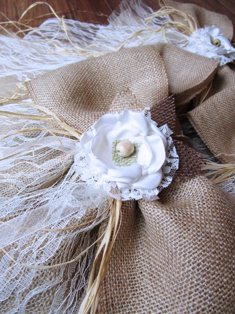 May include: A burlap and lace wedding ring pillow with a white fabric flower and a brown burlap bow.