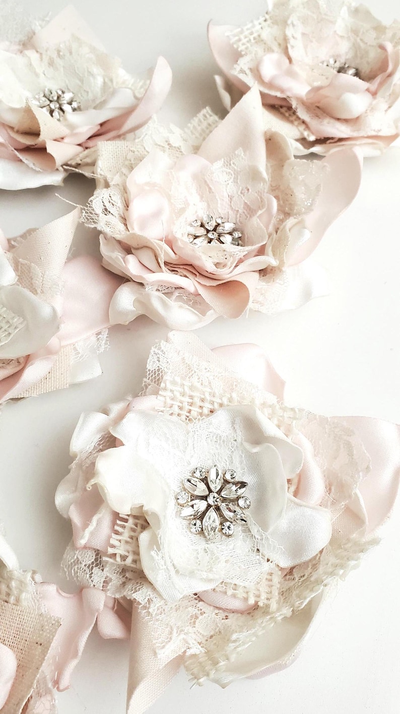 small fabric flowers made with bits of burlap, cotton, and lace. the center is accented by rhinestone small buttons. blush and ivory colors