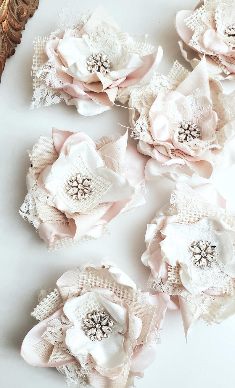 small fabric flowers made with bits of burlap, cotton, and lace. the center is accented by rhinestone small buttons. blush and ivory colors