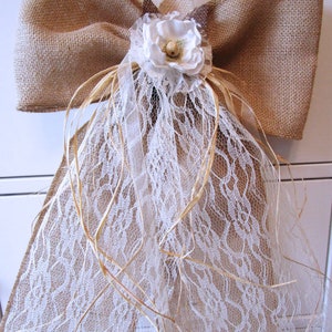 May include: A large burlap bow with a white lace overlay and a white fabric flower with a brown center. The bow is tied with twine and has a rustic, country feel.