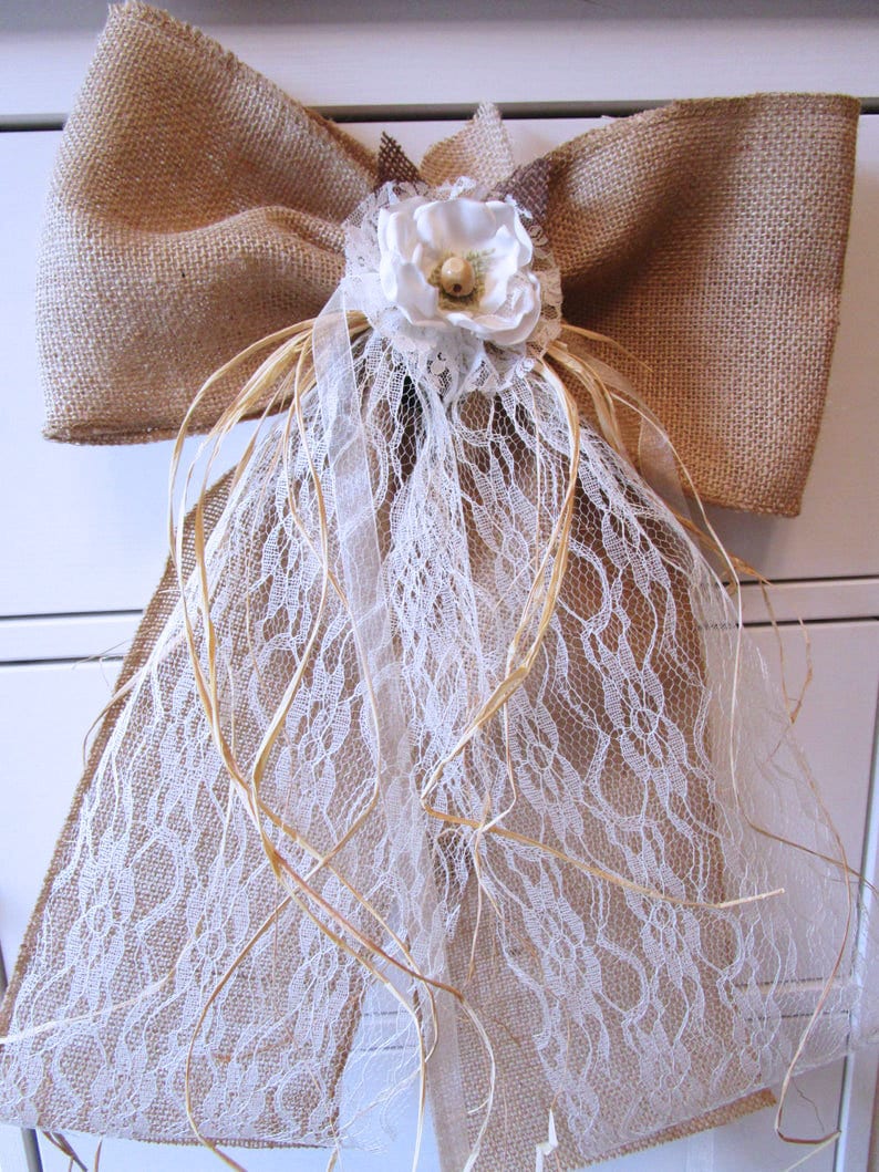 May include: A large burlap bow with a white lace overlay and a white fabric flower with a brown center. The bow is tied with twine and has a rustic, country feel.