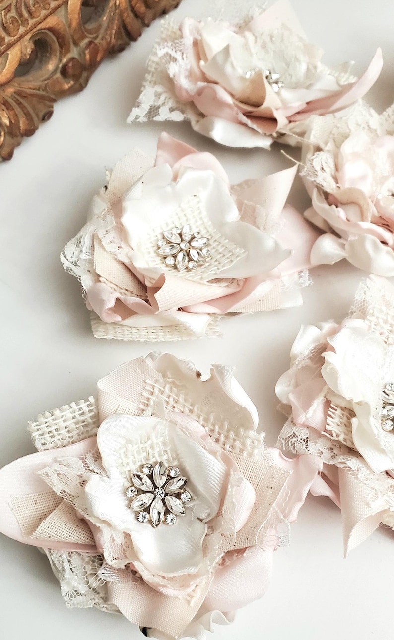 May include: A collection of handcrafted fabric flowers in shades of pale pink and cream. Each flower features layered satin and burlap petals, delicate white lace trim, and a central cluster of clear, faceted rhinestones arranged in a star shape.