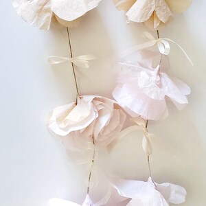 May include: A garland of delicate, handmade paper flowers in soft shades of pink and cream. The flowers are attached to thin, natural-colored twine with small ribbon accents, creating a romantic and airy aesthetic.