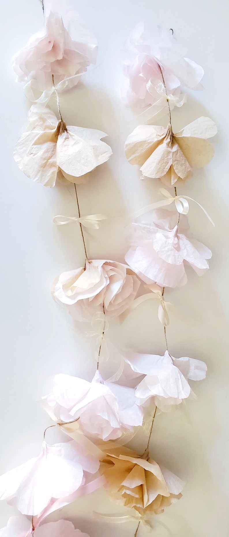 May include: A garland of delicate, handmade paper flowers in soft shades of pink and cream. The flowers are attached to thin, natural-colored twine with small ribbon accents, creating a romantic and airy aesthetic.