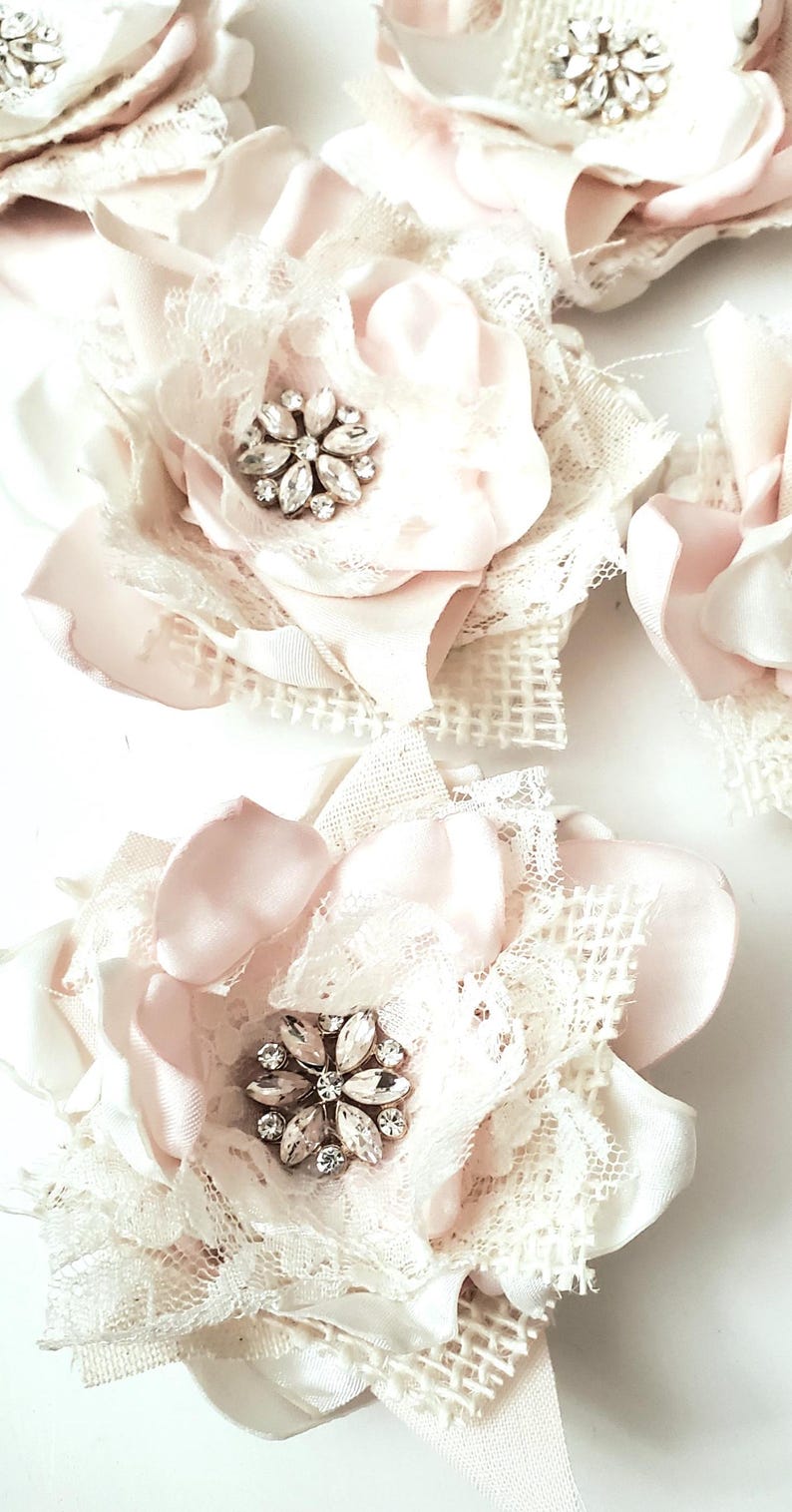 small fabric flowers made with bits of burlap, cotton, and lace. the center is accented by rhinestone small buttons. blush and ivory colors