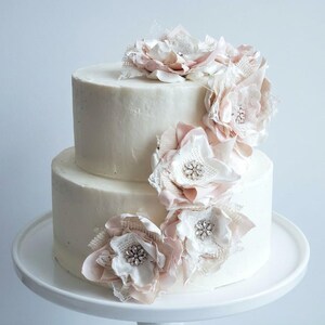 May include: A two-tier white frosted cake decorated with a cascading arrangement of fabric flowers in shades of pale pink and cream. Each flower features a central cluster of clear crystals and is accented with delicate lace and mesh fabric.