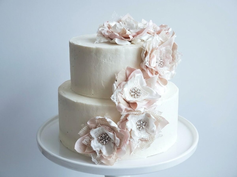 May include: A two-tier white frosted cake decorated with a cascading arrangement of fabric flowers in shades of pale pink and cream. Each flower features a central cluster of clear crystals and is accented with delicate lace and mesh fabric.
