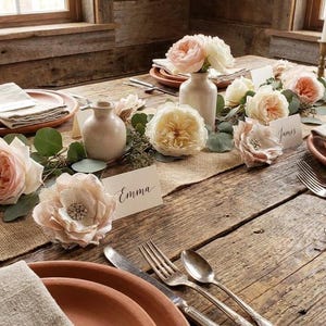 May include: A rustic table setting featuring terracotta plates, silver cutlery, and a centerpiece of cream and blush roses, eucalyptus greenery, and small ceramic vases. White place cards with the names Emma and James are placed on the table.