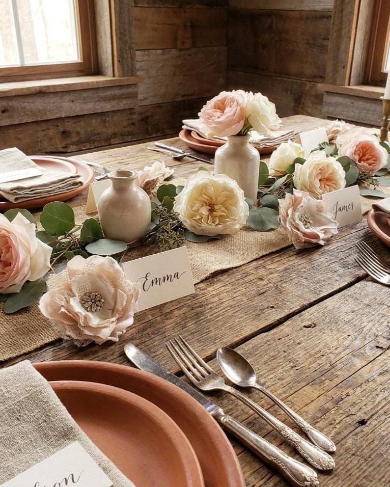 May include: A rustic table setting featuring terracotta plates, silver cutlery, and a centerpiece of cream and blush roses, eucalyptus greenery, and small ceramic vases. White place cards with the names Emma and James are placed on the table.