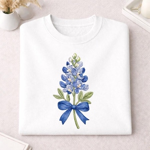 May include: White sweatshirt featuring a detailed illustration of a bluebonnet flower with a blue bow. The bluebonnet is a vibrant blue with green leaves and stem. The sweatshirt is made of a soft material.