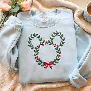 May include: A light blue sweatshirt featuring a Mickey Mouse head design made of green leaves, red berries, and a red bow. The sweatshirt is a crew neck style and is likely made of a soft material.