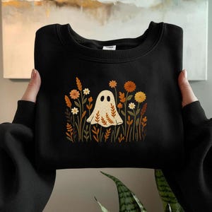 Puede incluir: Sudadera negra con un diseño de fantasma beige rodeado de flores y follaje de colores otoñales. El fantasma tiene dos ojos negros y un patrón de hojas en su cuerpo. El diseño está centrado en la parte delantera.