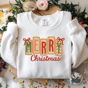 May include: White sweatshirt featuring the words "MERRY Christmas" in a festive, biscuit-style design. The letters are red, green, and pink, with red bows. The sweatshirt has a crew neck and long sleeves.