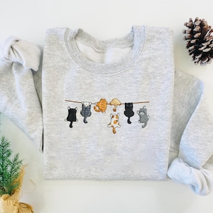 May include: A light gray sweatshirt featuring an embroidered design of six cartoon cats hanging from a brown line. The cats are in various colors, including black, gray, orange, and white. The sweatshirt has a crew neck and long sleeves.