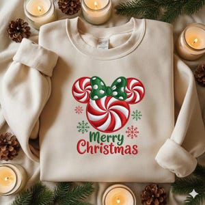 May include: Beige sweatshirt with a red and white candy cane design in the shape of Mickey Mouse ears, a green polka dot bow, and the words "Merry Christmas" in red and green. The sweatshirt has a crew neck and long sleeves.