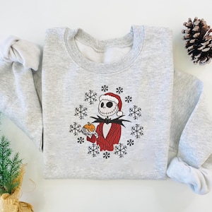 May include: A light gray sweatshirt featuring a festive design. The graphic depicts a skeleton character in a Santa hat and red suit, holding a pumpkin, surrounded by snowflakes. A pine cone and small evergreen tree are also visible.