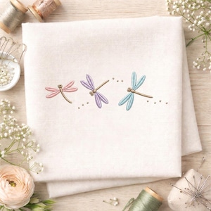 Op de afbeelding: Een witte linnen doek geborduurd met drie libellen in pastelkleuren: roze, paars en lichtblauw. De libellen zijn horizontaal gerangschikt met kleine geborduurde stippen erachter. De doek is gevouwen en heeft een zacht, getextureerd uiterlijk.