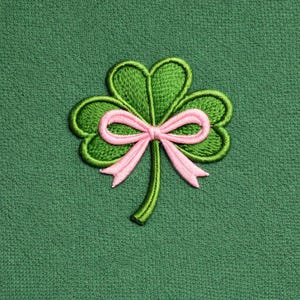以下が含まれることがあります： ピンクのリボンが付いた、緑色の刺繍されたクローバー。クローバーは、緑色の輪郭を持つ3つのハート型の葉を持っています。リボンはピンク色で、クローバーの中央で結ばれています。背景は無地の緑色の生地です。
