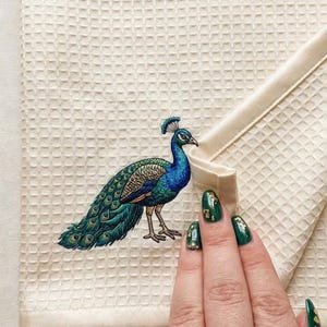 May include: Cream-colored waffle-weave towel featuring an embroidered peacock in shades of blue and green. The peacock has a detailed tail and crest. The towel has a folded edge.