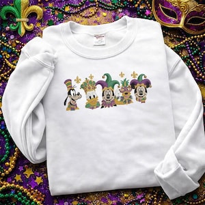 Puede incluir: Sudadera blanca con personajes de dibujos animados bordados con trajes de Mardi Gras. El diseño incluye a Goofy, el Pato Donald, Mickey Mouse y otros personajes con sombreros y coronas de bufón. La sudadera está rodeada de cuentas y una máscara de Mardi Gras.