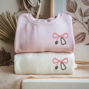 Peut inclure: Deux sweatshirts, un rose pâle et un crème, empilés. Chacun présente un motif brodé d'un nœud rose et d'un stéthoscope. Les sweatshirts sont pliés et reposent sur un tissu rose clair. Fond neutre.