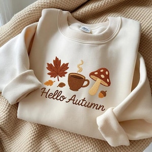 Può includere: Felpa color crema con ricamo a tema autunnale. Presenta una foglia d'acero marrone, una tazza di caffè, un fungo e la scritta "Hello Autumn" in marrone. Le maniche lunghe hanno polsini a coste.