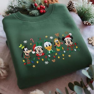 Puede incluir: Sudadera verde oscuro con bordados de personajes de Disney con atuendos navideños. El diseño incluye a Mickey, Minnie, Donald, Goofy y Pluto, con luces navideñas y bastones de caramelo.