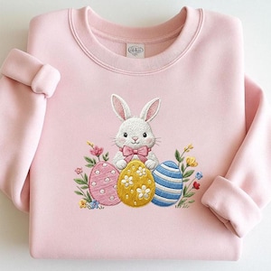 Op de afbeelding: Een lichtroze sweatshirt met een gedetailleerd borduurontwerp. Het ontwerp toont een witte konijn met een roze strik, drie versierde paaseieren en kleurrijke bloemen. De mouwen zijn opgerold.