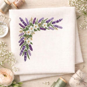 以下が含まれることがあります： 紫色のラベンダー、白い花、緑の葉をあしらったコーナー刺繍デザインの白いリネン布。 花の配置は詳細で繊細で、自然で植物的なテーマを作り出しています。 布は折りたたまれており、清潔で鮮明な外観を示しています。