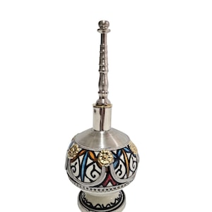 Può includere: Un flacone di profumo decorativo argentato con un lungo tappo ornato. Il corpo sferico presenta un design colorato con accenti dorati. La base è bianca con dettagli neri. Il testo "fezcraftswork" è visibile.