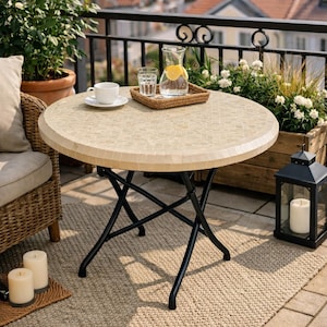 Op de afbeelding: Ronde tuintafel met een mozaïektegelblad en een opvouwbaar zwart metalen onderstel. Op de tafel staan een witte kop en schotel, een dienblad met een glas water en een karaf water met citroenschijfjes.