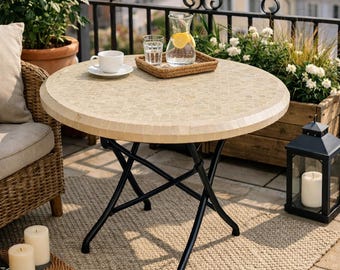Puede incluir: Mesa redonda de jardín con tablero de mosaico y base plegable de metal negro. La mesa está puesta con una taza y platillo blancos, una bandeja con un vaso de agua y una jarra de agua con rodajas de limón.