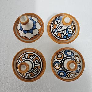Peut inclure: Quatre couvercles de tagine en céramique avec des motifs peints à la main. Chaque couvercle présente un motif unique en bleu, orange et marron sur fond blanc, avec un petit bouton central. Les couvercles ont une esthétique chaleureuse et terreuse.