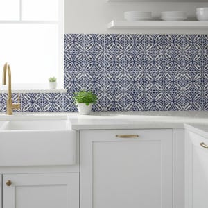 May include: A kitchen with white cabinets, a white sink, and a gold faucet. The backsplash features patterned blue and white tiles. A small potted plant sits on the countertop, and another is on the windowsill.