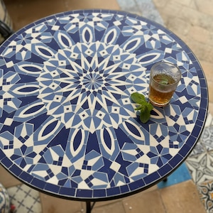 May include: A round table featuring a mosaic top with a geometric pattern in shades of dark blue, light blue, and white. A clear glass containing amber-colored liquid and a sprig of fresh green mint leaves sits on the surface of the table.