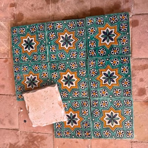Puede incluir: Un conjunto de ocho azulejos cuadrados de cerámica con fondo verde azulado y un patrón central de estrella de ocho puntas en amarillo y blanco con un motivo floral azul oscuro, rodeado de pequeños detalles florales blancos y naranjas.