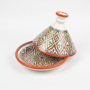 Peut inclure: Un tajine en céramique avec un couvercle conique et une base ronde. Le tajine présente un motif vibrant de motifs géométriques verts, rouges et blancs sur fond blanc. Le bord et le dessus du couvercle sont orange.