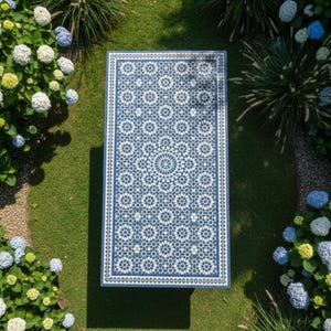 Pode incluir: Uma mesa retangular ao ar livre com um padrão geométrico azul e branco. A mesa é cercada por grama verde e flores coloridas. O design da mesa apresenta um medalhão central e formas de estrela repetidas.