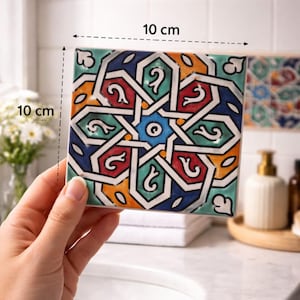 Op de afbeelding: Vierkante keramische tegel met een kleurrijk geometrisch patroon. De tegel is 10 cm x 10 cm en heeft een stervormig ontwerp in blauw, rood, oranje en groen, met witte contouren. De tegel wordt voor een badkameromgeving gehouden.