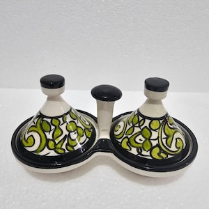 Może przedstawiać: Ceramiczny serwer do przypraw z dwoma komorami w kształcie miski i centralnym uchwytem. Miski zdobione są zielono-białymi wzorami kwiatowymi i czarnymi obwódkami. Pokrywki i uchwyt są czarne.