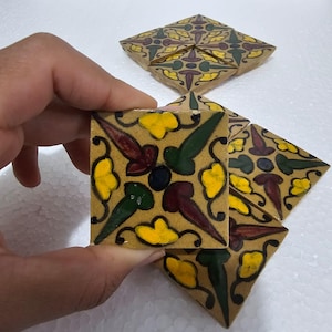 Peut inclure: Un carreau de carrelage carré de couleur beige, orné d'un motif floral jaune, vert et marron, est tenu par une main. D'autres carreaux similaires sont visibles en arrière-plan, suggérant un ensemble de carrelage décoratif.