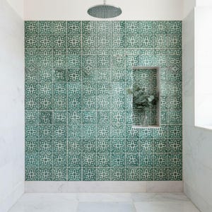 Può includere: Una parete doccia rivestita con piastrelle quadrate caratterizzate da un motivo floreale ripetitivo verde e bianco. Una nicchia rettangolare è integrata nella parete e contiene un vaso di vetro con fogliame verde. Un soffione circolare è montato sopra.