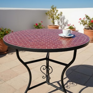 Könnte beinhalten: Ein runder Gartentisch mit einer Mosaik-Tischplatte in Rot- und Brauntönen. Der Tisch hat ein schwarzes Metallgestell mit dekorativen Schnörkeln. Eine weiße Kaffeetasse mit Untertasse steht auf dem Tisch. Topfpflanzen umgeben den Tisch.