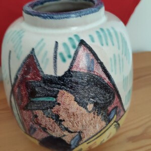 May include: A round, white ceramic vase with a blue rim and a colorful abstract design. The design features a face with black hair, red and brown accents, and green vertical lines. The vase sits on a wooden surface.