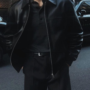 May include: A black leather jacket with a pointed collar, shoulder epaulets, and a full-length silver zipper, worn over a dark gray polo shirt and black trousers with a black belt featuring a silver buckle, creating a monochromatic ensemble.