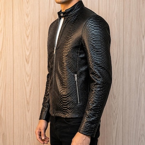 May include: A black jacket featuring a textured snakeskin pattern, a stand-up collar with a single snap button closure, a full-length silver-toned front zipper, and a zippered side pocket. The garment is worn over a white shirt and black pants.